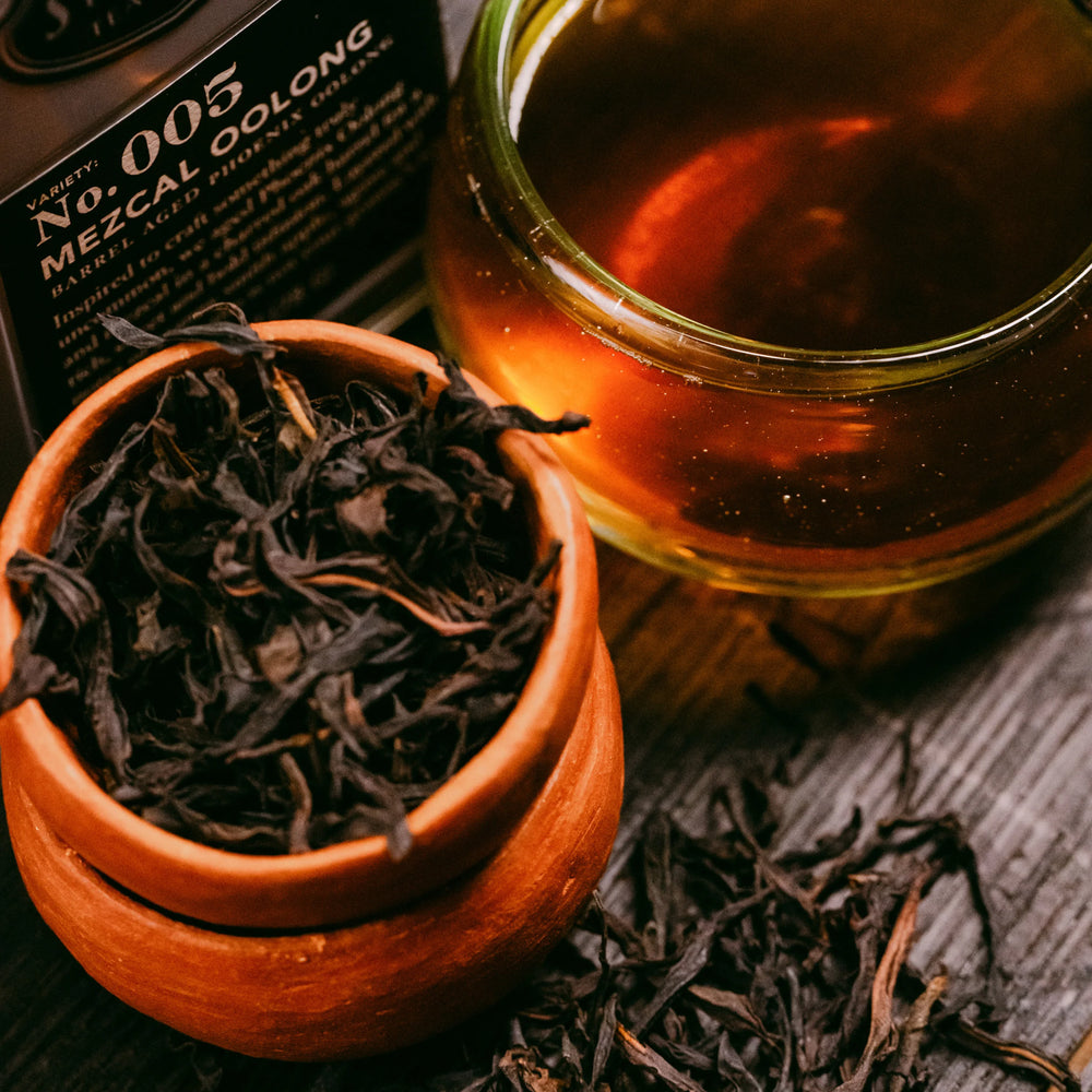 Tea leaves in a terracotta pot with a glass of tea and a bottle labeled 'No. 005 Mezcal Oolong' on a wooden surface.