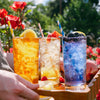 Three tall glasses of colorful iced drinks on a wooden tray outdoors, with a hand holding the tray. The drinks are amber, clear with lemon and raspberries, and blue-purple with blackberries, each with a striped straw.