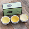 Green box with black trim on wooden table next to three porcelain cups filled with green teas.